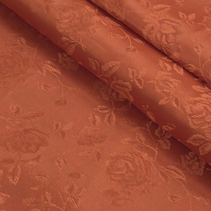 Puede incluir: Primer plano de una tela doblada con un patrón de rosas. La tela es de un cálido color terracota, con el diseño de rosas sutilmente en relieve. Los pliegues crean sombras, realzando la textura y la profundidad del motivo floral. La tela parece ser un material lujoso.