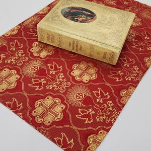 Op de afbeelding: Een rode stof met een gouden patroon van duiven en kruisen. Een goudkleurig boek met de tekst "Holy Bible" en "New Standard Reference Bible" ligt op de stof.