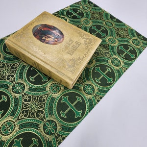 May include: A vintage Holy Bible book with a gold-colored cover and a green and gold patterned fabric underneath. The cover features a religious scene with people and a cross.