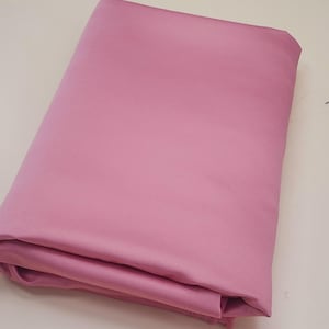 May include: A folded, solid pink fabric, likely a textile or material for crafting or sewing. The fabric has a smooth, even texture and a soft, muted pink color. The edges are neatly folded, suggesting it is a new, unused piece of fabric.