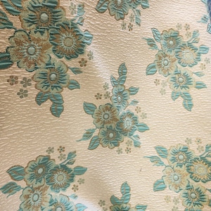 Puede incluir: Primer plano de una tela blanca con un patrón repetitivo de flores azul turquesa y doradas. La tela tiene una superficie texturizada.