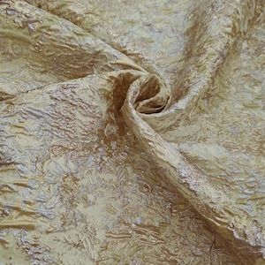 Puede incluir: Primer plano de una tela texturizada en tonos dorados y beige. El material presenta un patrón irregular en relieve, creando una sensación de profundidad. La tela está doblada y drapeada, mostrando su textura y brillo. Ideal para ropa o decoración del hogar.