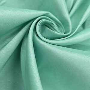 May include: Close-up of a folded, light-green fabric with a silky sheen. The fabric is arranged in soft folds and curves, creating a sense of depth and texture. The color is a muted, pastel green, reminiscent of seafoam or mint.