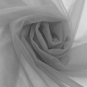 Puede incluir: Una tela de tul gris claro transparente con una estructura de malla hexagonal fina. El material está recogido en un suave patrón de espiral, creando capas superpuestas que varían en opacidad y resaltan la textura ligera y delicada.