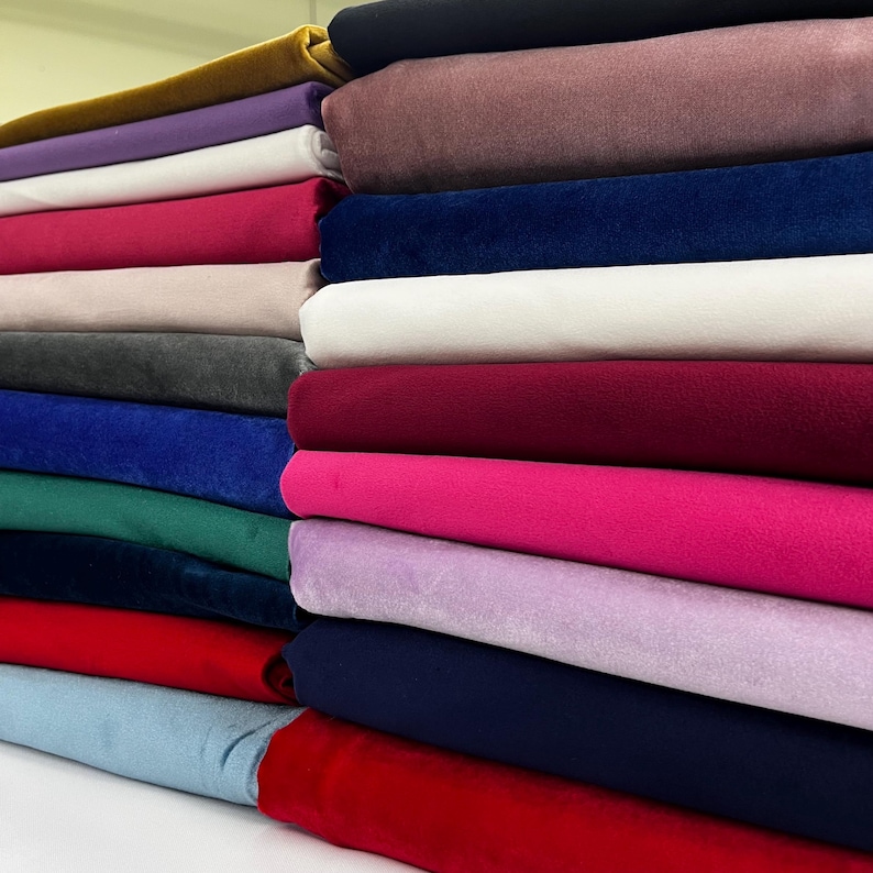 May include: A stack of various colored velvet fabric rolls. Colors include gold, purple, white, pink, gray, blue, green, red, and black. The fabric appears soft and plush, suitable for upholstery or crafting projects.