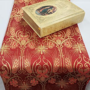 May include: A red and gold patterned textile with a book on top. The textile features a repeating design of crosses, wheat, and floral elements. The book has a gold cover with an oval image and the text "BIBLIA ZELATORUM".