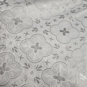May include: A white fabric with a silver metallic pattern of repeating quatrefoils and crosses. The pattern is embossed and has a subtle sheen.