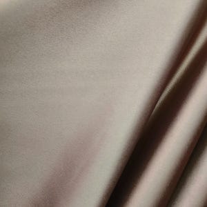 May include: A close-up of a smooth, light brown fabric with a subtle sheen. The fabric is folded in a way that creates a soft, flowing drape.