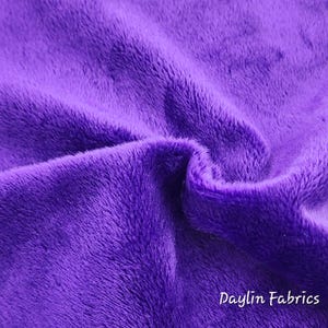 May include: Close-up of a soft, purple fabric with a fuzzy texture. The fabric is folded and draped, showing its plushness. The text "Daylin Fabrics" is visible in the bottom right corner.