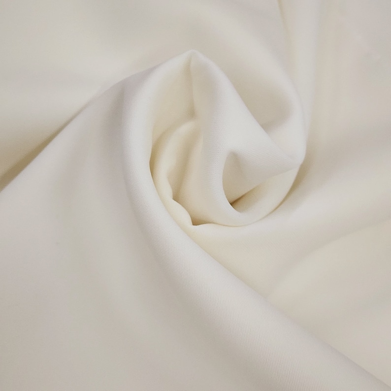 May include: Close-up of a cream-colored fabric with soft folds and subtle shadows. The fabric appears smooth and drapes gently, suggesting a lightweight material. The overall tone is neutral and elegant, suitable for apparel or home decor.