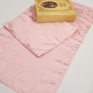 May include: A pink satin prayer rug with a paisley pattern. A gold book with the words "Holy Bible" on the cover is on top of the rug.