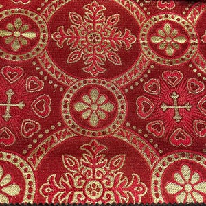 Puede incluir: Primer plano de una tela roja con un patrón dorado ornamentado. El diseño incluye motivos florales, de cruz y de corazón dentro de formas circulares y de copo de nieve. La tela tiene una apariencia texturizada.
