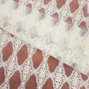 May include: Ivory lace fabric with a floral pattern. The lace is layered over a brown fabric, creating a contrast in color and texture. The floral design is repeated across the fabric.