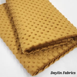 May include: A close-up of a folded piece of mustard yellow minky fabric with a textured dot pattern. The fabric is soft and plush.