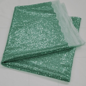 Op de afbeelding: Een tealgroene paillettenstof met een transparante overlay. De stof is dubbelgevouwen en de pailletten zijn aan beide kanten zichtbaar.