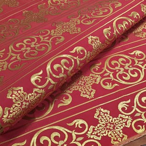 Puede incluir: Tela roja con adornos dorados de volutas y motivos florales. La tela está doblada, revelando el diseño repetitivo. Los elementos dorados están en relieve, creando un efecto texturizado. Adecuado para tapicería o proyectos decorativos.