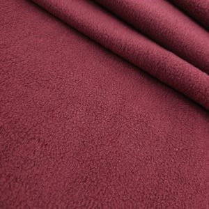 Puede incluir: Primer plano de una tela de forro polar burdeos. La tela es suave y texturizada, con pliegues visibles y un aspecto afelpado. El color es un tono profundo y rico de rojo-púrpura.