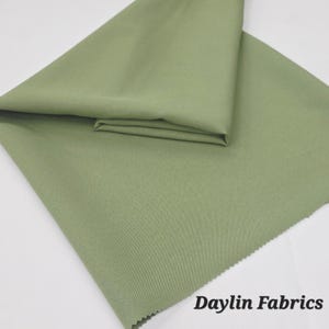 Puede incluir: Un trozo de tela verde salvia doblado. La tela es lisa y tiene una textura sutil. El texto "Daylin Fabrics" es visible en la esquina inferior derecha de la imagen.