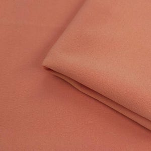 Puede incluir: Primer plano de una tela doblada de color melocotón. La tela tiene un aspecto suave y texturizado, con sutiles variaciones de tono. Los pliegues crean sombras, añadiendo profundidad a la imagen. La tela parece ser un material ligero.