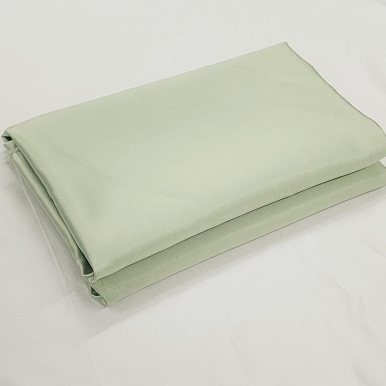 May include: Folded light green fabric, possibly a sheet or tablecloth, with a smooth, slightly shiny texture. The fabric is neatly folded, showing a clean edge and a soft, muted color. The fabric is likely made of a lightweight material.