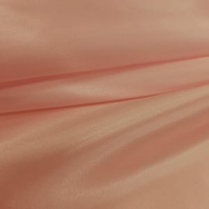 Puede incluir: Primer plano de una tela color melocotón con brillo satinado. El material está doblado, creando sombras suaves y resaltando la textura lisa. La tela parece ligera y fluida, adecuada para prendas de vestir o fines decorativos.