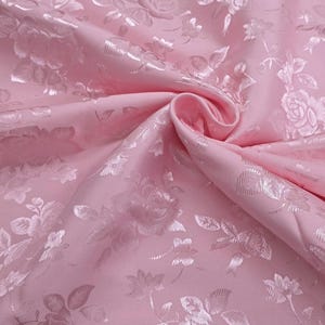 May include: Pink fabric with a floral pattern. The fabric has a satin sheen and a raised floral design. The roses and leaves are in a lighter shade of pink, creating a subtle contrast. The fabric is folded, showing the texture and depth.