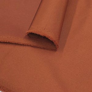 Puede incluir: Primer plano de una sección de tela doblada de color óxido. La tela tiene una textura suave y un ligero brillo. Los bordes de la tela están deshilachados. Las palabras "Daylin Fabrics" están impresas en blanco en la parte inferior derecha.