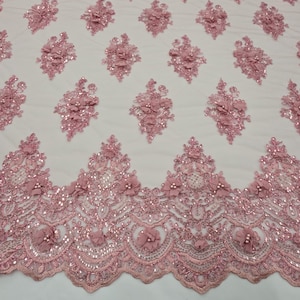 Puede incluir: Tela de encaje rosa con adornos florales y lentejuelas. La tela presenta un patrón repetitivo de flores 3D y lentejuelas, con un borde festoneado. El encaje es de color rosa suave y las flores tienen un aspecto delicado y texturizado.