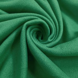 Puede incluir: Primer plano de una tela verde vibrante con una superficie suave y texturizada. El material está enrollado, creando un patrón arremolinado. El color es un verde rico y sólido, lo que sugiere un textil adecuado para diversos proyectos de artesanía o ropa.