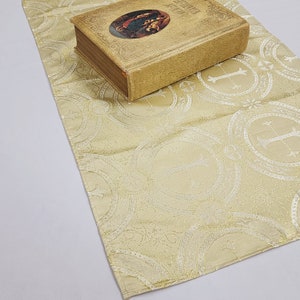 以下が含まれることがあります： 装飾的な枠の中に白い十字架が繰り返し描かれた、ベージュの布製の祭壇布。表紙が金色で、宗教的な絵が描かれた閉じた本が布の上に置かれている。