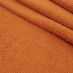 Puede incluir: Primer plano de una tela de forro polar naranja doblada. La tela tiene una superficie suave y texturizada y está dispuesta en pliegues diagonales, creando sombras y profundidad. El color es un tono cálido y acogedor de naranja.