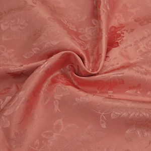 May include: Close-up of a peach-coloured fabric with a floral pattern. The fabric has a satin sheen and a raised, embossed design of roses and leaves. The folds in the fabric create shadows and highlights, adding depth to the image.