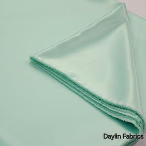 Pode incluir: Close-up de uma pilha de tecido de cetim verde menta. O tecido tem uma superfície lisa e brilhante e uma borda macia e texturizada. As palavras "Daylin Fabrics" estão impressas em branco no canto inferior direito.