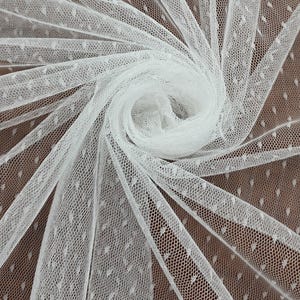 Puede incluir: Primer plano de tela de tul blanca con un delicado estampado de lunares. La tela está dispuesta en espiral, mostrando su textura y transparencia. El fondo es de un marrón cálido, que contrasta con el material blanco brillante.