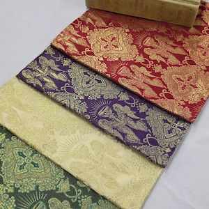 May include: A selection of patterned fabrics in red, purple, cream, and green, with gold angel and floral designs. A vintage book is placed on top. The fabrics are folded and arranged at an angle, showcasing the intricate patterns.