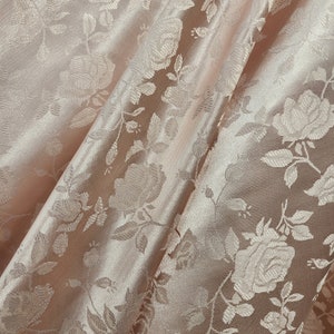 Könnte beinhalten: Nahaufnahme eines hellrosa Stoffes mit einem erhabenen Blumenmuster. Der Stoff hat einen satinartigen Glanz und ist weich im Griff.