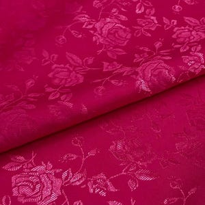 Könnte beinhalten: Nahaufnahme eines leuchtend pinkfarbenen Stoffes mit einem floralen Rosenmuster. Der Stoff ist gefaltet und zeigt die Textur und die komplizierten Details der geprägten Rosen und Blätter. Das Material hat einen dezenten Glanz.
