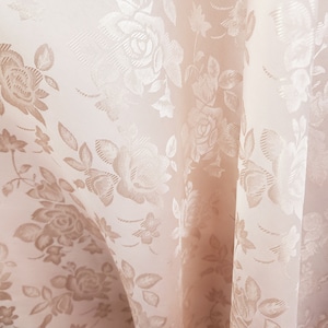 Könnte beinhalten: Nahaufnahme eines blassrosa Stoffes mit einem zarten, geprägten Blumenmuster. Der Stoff hat einen dezenten Glanz und eine weiche, fließende Textur.