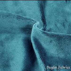 May include: Close-up of teal-colored plush fabric with a soft, textured surface. The fabric is folded, revealing the depth and texture of the material. The Daylin Fabrics logo is visible in the bottom right corner.