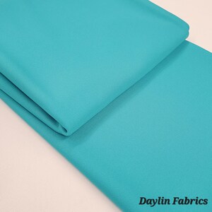 May include: A folded piece of turquoise blue fabric. The fabric is smooth and has a slight sheen. The fabric is folded in half and the bottom edge is visible. The fabric is on a white surface. Daylin Fabrics is printed in black text at the bottom right corner of the image.