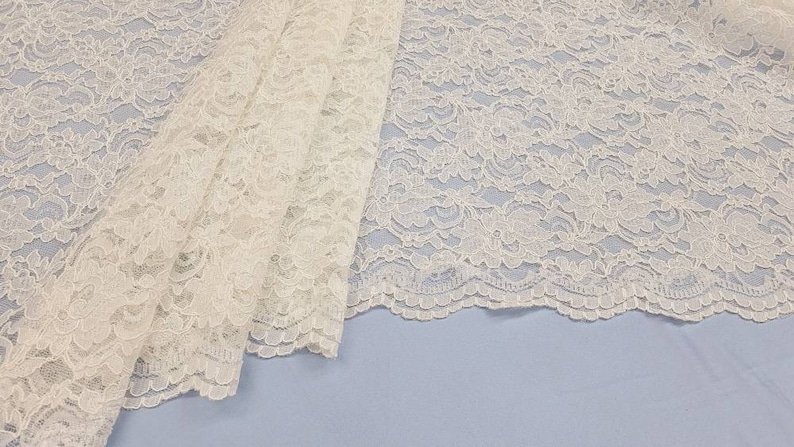May include: White lace fabric with a delicate floral pattern. The lace has a scalloped edge.