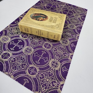 May include: A purple fabric with a gold pattern of crosses and circles. A gold book with the title "Holy Bible" is on top of the fabric.