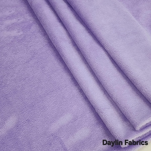 Può includere: Primo piano di un tessuto morbido di colore lilla con una texture soffice. Il tessuto è piegato in più strati, creando un aspetto liscio e fluido. Il testo "Daylin Fabrics" è visibile nell'angolo in basso a destra dell'immagine.