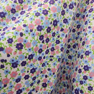 May include: White fabric with a repeating pattern of small pink, blue, and purple flowers. The fabric has a subtle texture and a slightly wrinkled appearance.
