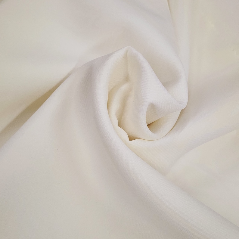 May include: Close-up of a cream-colored fabric with soft folds and a smooth texture. The fabric is tightly bunched in the center, creating a swirling pattern. The material appears to be lightweight and flowing, ideal for apparel or home decor.