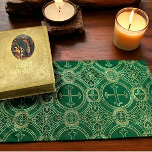 May include: A green fabric altar cloth with a gold pattern of crosses and circles. A gold-colored book with the words "Holy Bible" on the cover is on top of the cloth. Two lit candles are in the background.