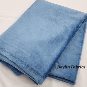 Może przedstawiać: Złożony kawałek miękkiej, jasnoniebieskiej tkaniny polarowej. Tkanina jest gładka i ma delikatny połysk. Tekst "Daylin Fabrics" jest nadrukowany na biało w prawym dolnym rogu tkaniny.