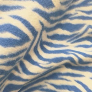 Puede incluir: Una tela suave y difusa que presenta un patrón repetitivo de rayas de cebra en tonos azul claro y blanco roto. El material tiene una textura densa y afelpada con pliegues y ondulaciones visibles que crean profundidad en la superficie.