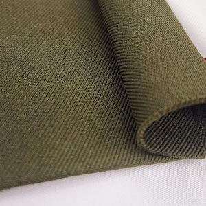 Op de afbeelding: Close-up van een groene stof met een getextureerd, geribbeld oppervlak. De stof is gevouwen en de randen zijn zichtbaar.
