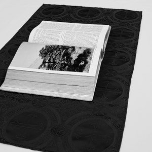 May include: An open book with a black and white photograph rests on a black fabric runner. The runner features a repeating pattern of crosses within circles. The book's pages are filled with text.